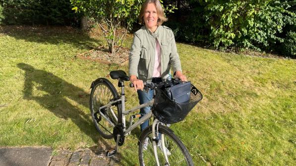 A woman is standing with her hybrid bike. She is wearing blue jeans, trainers and a pale green jacket. Her bike has a basket on the front with her handbag in it. She has blonde hair and is smiling at the camera