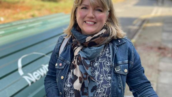 A woman is smiling into the camera. She is holding a bike and is wearing a blue denim jacket, multicoloured scarf and blue shirt with a white flower pattern. Behind her is an on-street cycle hub.
