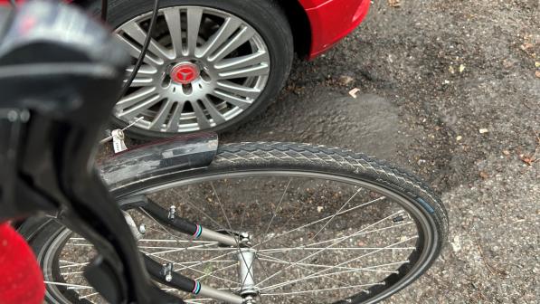 A large pothole in an urban street with a red car's wheel in it and a black bike wheel next to it