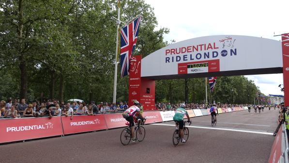 People are riding on a wide road towards a finish line that reads 'Prudential RideLondon' and has the time. The road is lined with spectators