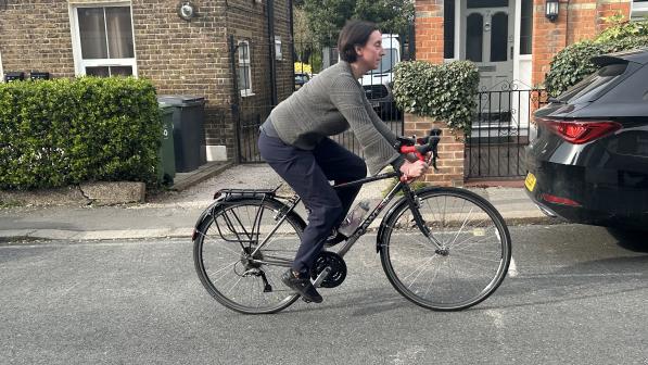 A woman is cycling a black touring bike on an urban road with houses and parked cars in the background. She's wearing a green sweater and blue trousers.