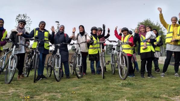 A big group of women is standing on a field all holding bikes. They are all wearing headscarves and are smiling. One is holding a baby. They are waving at the camera