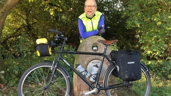 A man in cycling kit and glasses is leaning on a stone marker with a metal ball in the top with a metal plaque below which has Neptune written on it. A loaded touring bike is leaning against the marker