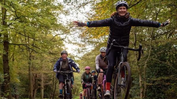 A group of women are riding their bikes on a muddy track through a forest. The woman at the front is standing up in her pedals and has her arms out to the side in a a gesture of celebration