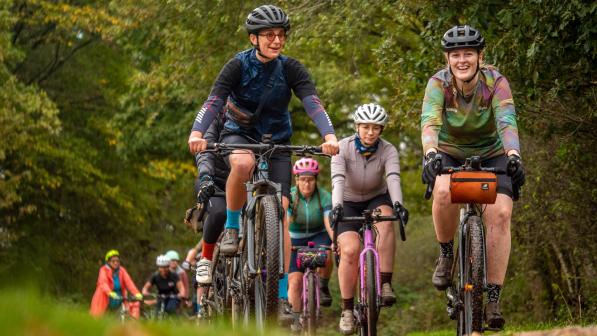 A group of women are cycling in a muddy forest. They're wearing mountain biking kit and on a mix of gravel and mountain bikes