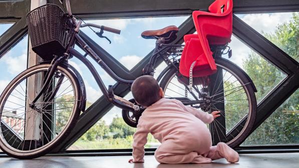 A toddler in a pink babygro is crawling towards a black Dutch-style bike with a red child seat on the back