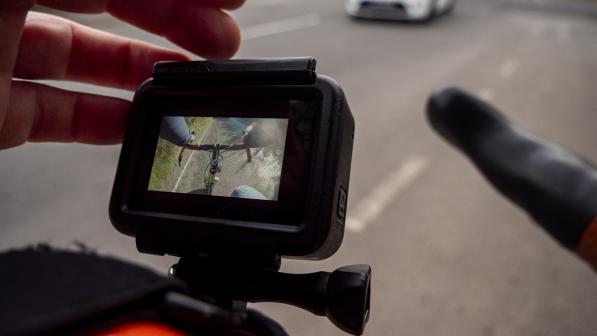 A close-up of a someone adjusting a helmet cam for a cycling helmet, the orange helmet can just be seen. The camera screen shows the bike's handlebar