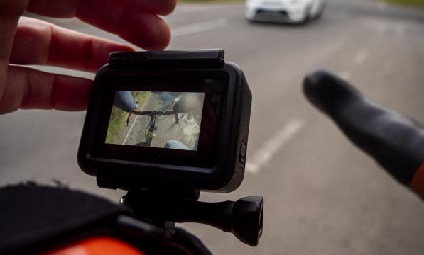 A close-up of a someone adjusting a helmet cam for a cycling helmet, the orange helmet can just be seen. The camera screen shows the bike's handlebar