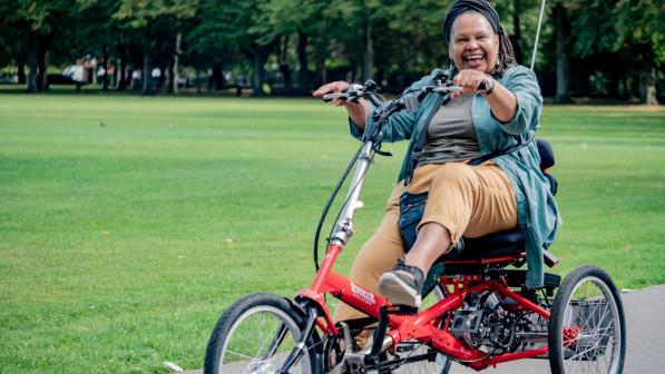 A smiling adult rides a red recumbent trike on a park path, one hand lifted, with trees and grass in the background.