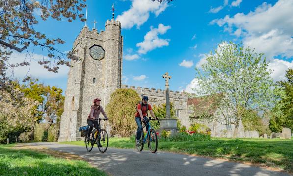 Two people on touring bikes with luggage are cycling past a country church in spring