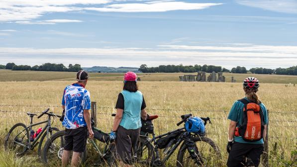 A group of three people with loaded touring bikes are standing looking out at Stone Henge in the background
