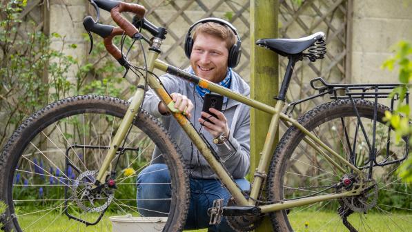 A man is cleaning a very dirty mountain bike. He's crouching down and has a cloth in one hand and is cleaning the down tube. In the other hand he's holding a smartphone and is wearing headphones. He's smiling.