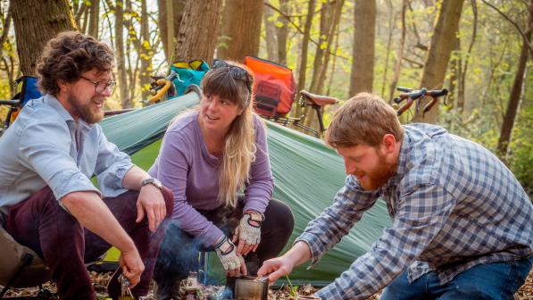 Three people are camping in the woods. They are making a hot drink. There's a tent in the background and you can see two packed gravel bikes leaning on trees