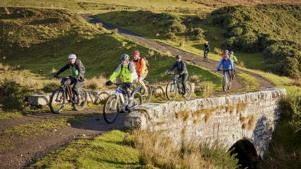A group of people are cycling over an old stone bridge on an off-road track through the Highlands. It's autumn and they're wearing warm cycling kit.
