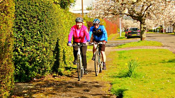Two cyclists, one in a bright pink jacket and the other in a blue jacket, ride along a scenic, tree-lined path adorned with blooming cherry blossoms. The vibrant greenery and spring flowers create a picturesque and inviting atmosphere, perfect for enjoying a leisurely cycling experience.