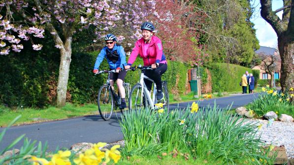 Two people cycling along a scenic, flower-lined path under blooming trees, enjoying the fresh air and vibrant colours of spring.