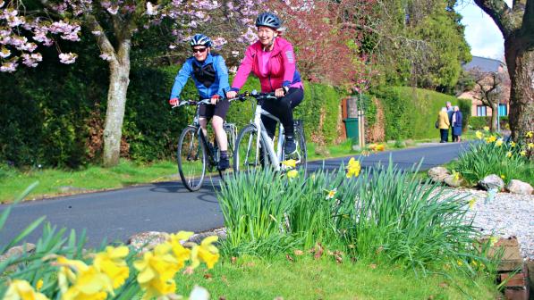 Two people cycling along a scenic, flower-lined path under blooming trees, enjoying the fresh air and vibrant colours of spring.