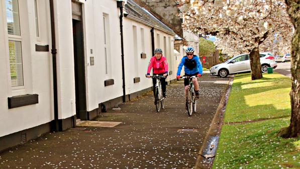 Two people cycling along a scenic, flower-lined path under blooming trees, enjoying the fresh air and vibrant colours of spring.