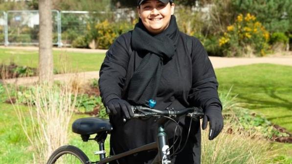 A woman is standing astride holding a bike in a park. She is wearing a black scarf and long black dress, as well as a black cap. Her bike is a dark metallic green hybrid. She is smiling at the camera. Photo: Sonny Malhotra