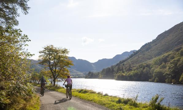 Two people cycling the Traws Eryri route, next to a lake with mountains and sunshine