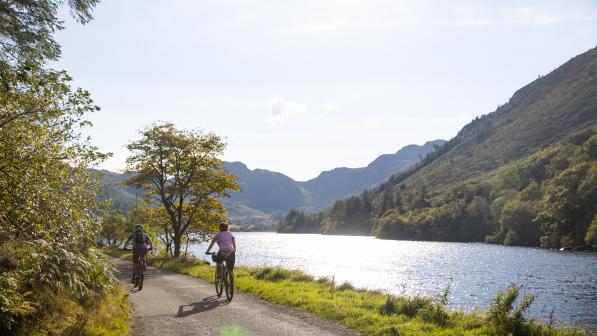 Two people are cycling on an off-road track next to a lake. They are on mountain bikes, and are both wearing shorts and T-shirts. There are mountains in the background.