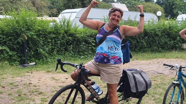 ALT TEXT: Sara Ramsey is sitting astride a black Trek touring bike. She is wearing shorts and a T-shirt. She has her arms up in ‘strong man’ pose and she’s smiling at the camera.