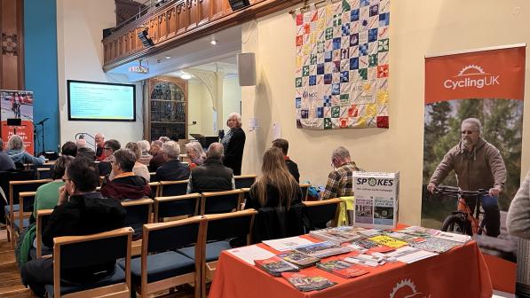 A group of people is sitting in chairs facing a stage. Behind them is a table with an orange tablecloth with the Cycling UK logo on it and lots of flyers and information on the table. There's also a poster featuring a man on a bicycle.