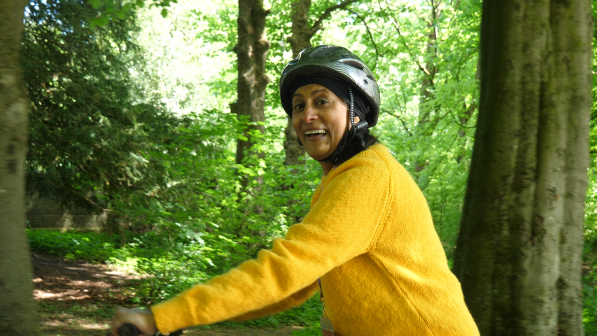 A woman wearing a yellow jumper and a helmet smiles while cycling through a wooded area, surrounded by tall trees and greenery.