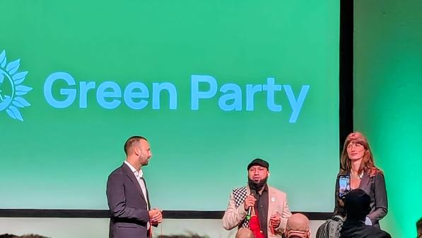 Three people are standing on a stage, with one in middle holding a microphone as he introduces the other two, a man and a woman. Behind them is a big green screen that reads 'Green Party'