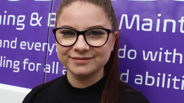 A head and shoulders shot of Sophie Bujdoso. She has long brown hair pulled back in a ponytail and is wearing glasses and a black sweater. She is smiling into the camera. She’s standing in front of the back of a van 