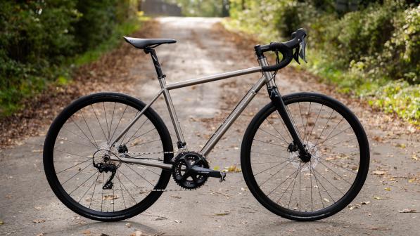 The Spa Cycles Elan Ti Mk2 105 12-speed Hydraulic, a silver touring bike, is propped up on a tarmac, leaf-strewn road with trees and hedges lining both sides