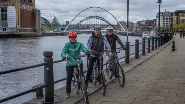 Three people are standing astride their bikes next to the River Tyne. The BALTIC and Sage and the Tyne bridges can be seen in the background