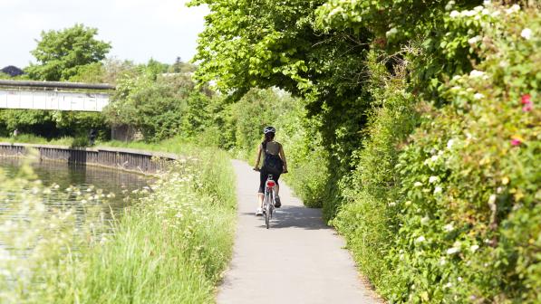 A woman is riding along a paved path next to a canal with a line of trees to the other side. It's sunny, she's wearing summer clothes and is cycling away from the camera