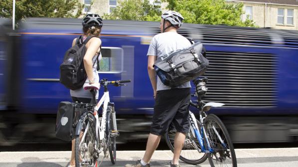 Two people with bikes are standing on a railway platform with a train going past them. They have packed rucksacks and bags and are wearing summer clothes and cycle helmets