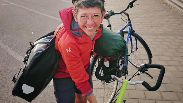 Susanna Thornton is looking up at the camera while holding onto a bike. She has two packed panniers slung across her shoulders. She is wearing shorts and a jacket and glasses; she also has sunglasses on her head. She is smiling.
