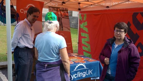 Three people are standing underneath an orange gazebo next to a table with a blue cloth on it that has the Cycling UK logo on