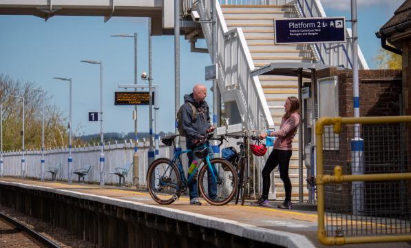 Two people are standing on a railway platform. They are both holding bikes.