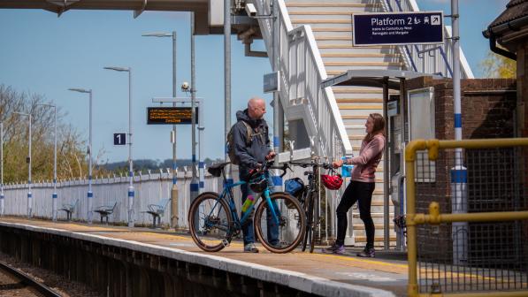 Two people are standing on a railway platform. They are both holding bikes.