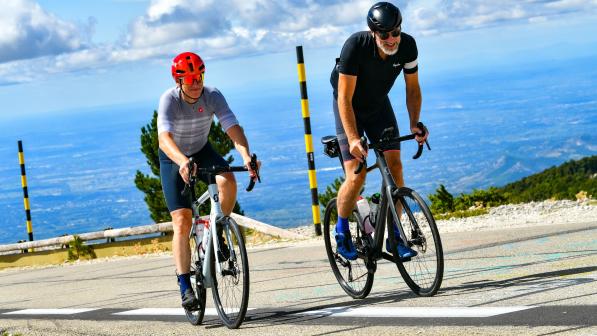 Two men on road bikes and wearing cycling kit and helmets are riding up a steep tarmac road