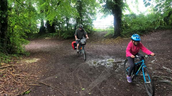 Two people are cycling through a muddy forest: a young girl on a blue mountain bike is in front and she's followed by a man on a flat-bar touring bike with packed pannier and bar bag.