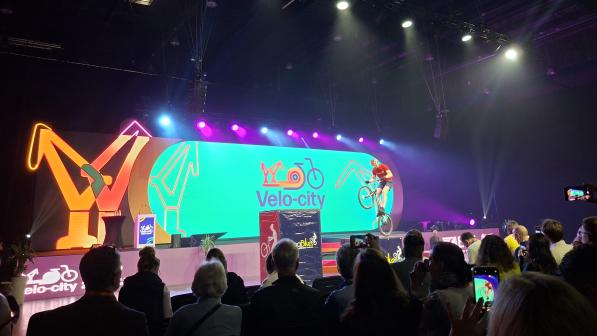 People are attending a conference. A person is doing tricks on a bike on the stage. The background of the stage is lit up turquoise panel that reads 'Velo-city' and has a bike design on it