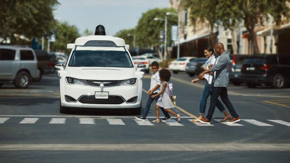 A white driverless car is stopped at a zebra crossing with a family of four crossing in front of it in a car-lined urban street