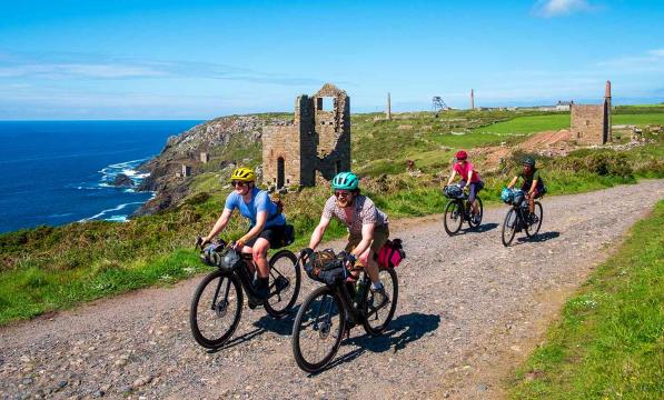 Four people cycling gravel bikes along the West Kernow Way, with a castle ruin and the sea in the background