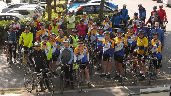 A big group of people are all standing on a pavement with their bikes. Most are wearing a Wheel Easy cycling jersey