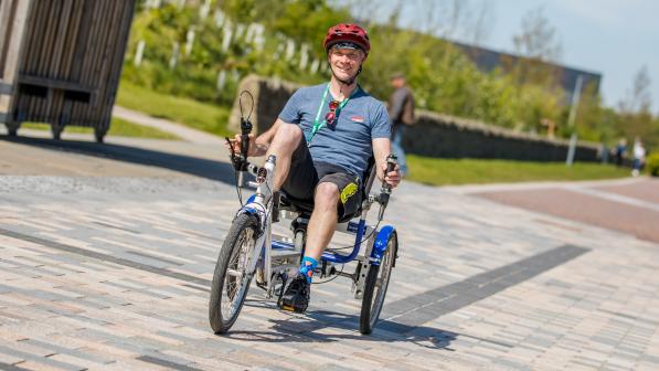 A man smiling at the camera as he cycles a recumbent trike out in the sun on a pedestrianised path