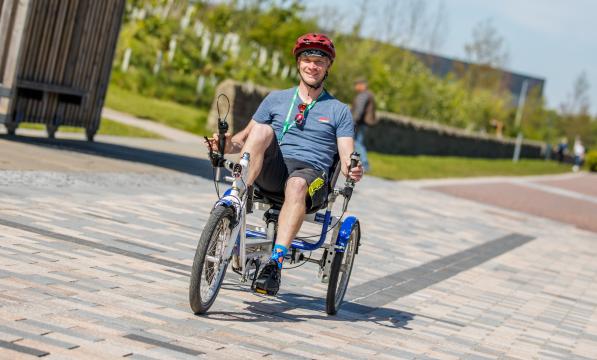 A man smiling at the camera as he cycles a recumbent trike out in the sun on a pedestrianised path