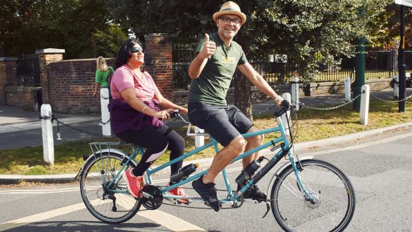 Two people are on a turquoise tandem cycling across a quiet junction with trees and a park in the background. The pilot is a man in shorts, T-shirt and sunhat. He's giving a thumbs up. The stoker is a woman in dark glasses. She's wearing leggings and a T-shirt.