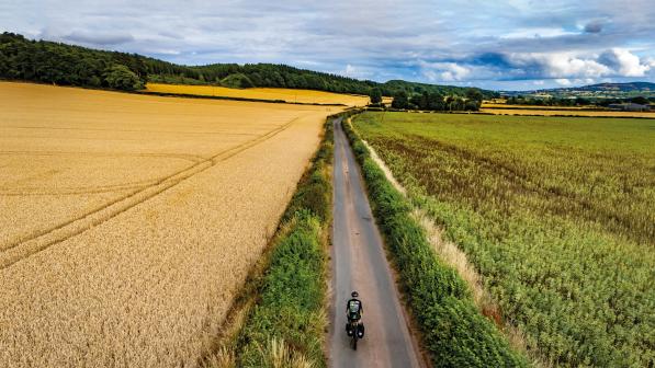A person is cycling along a straight singletrack road between two crop fields