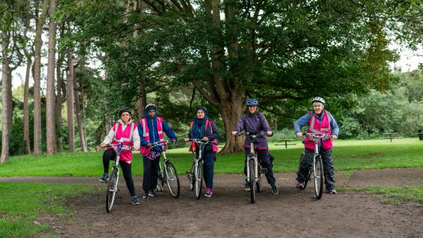 Five women are in a park standing with their bikes in front of a big tree. They are wearing a mix of cycling and normal clothing.
