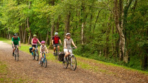 A group of people are riding along a leaf-littered dirt track through a forest. There are two women, two men and a little girl.